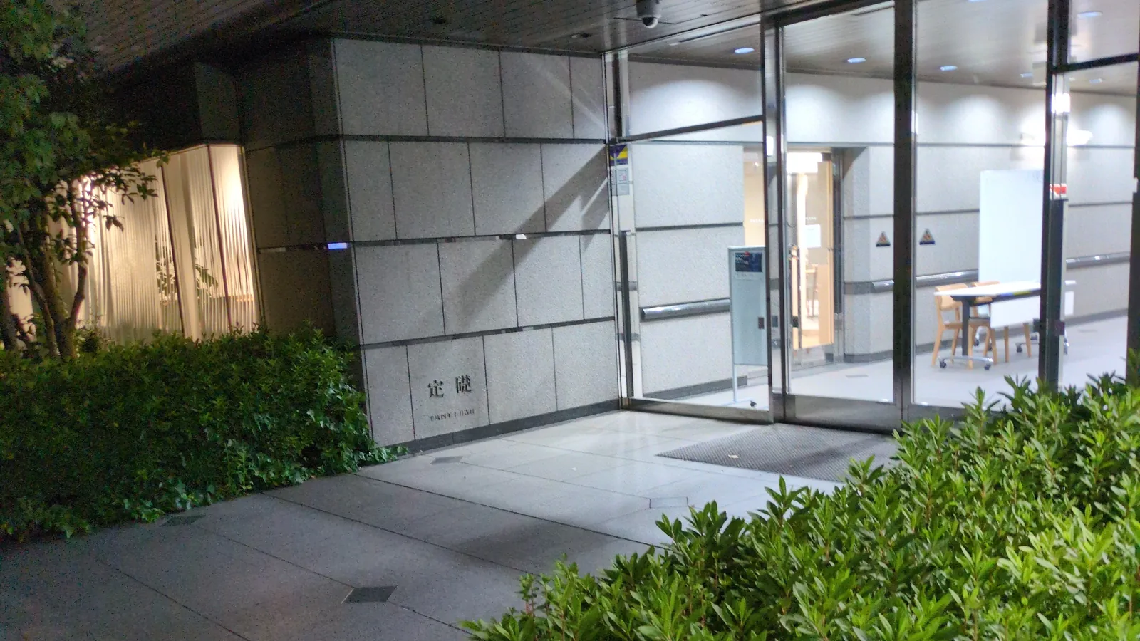 夜の会場入口。白い壁とガラス扉が照明に浮かび、左右を植え込みが囲む