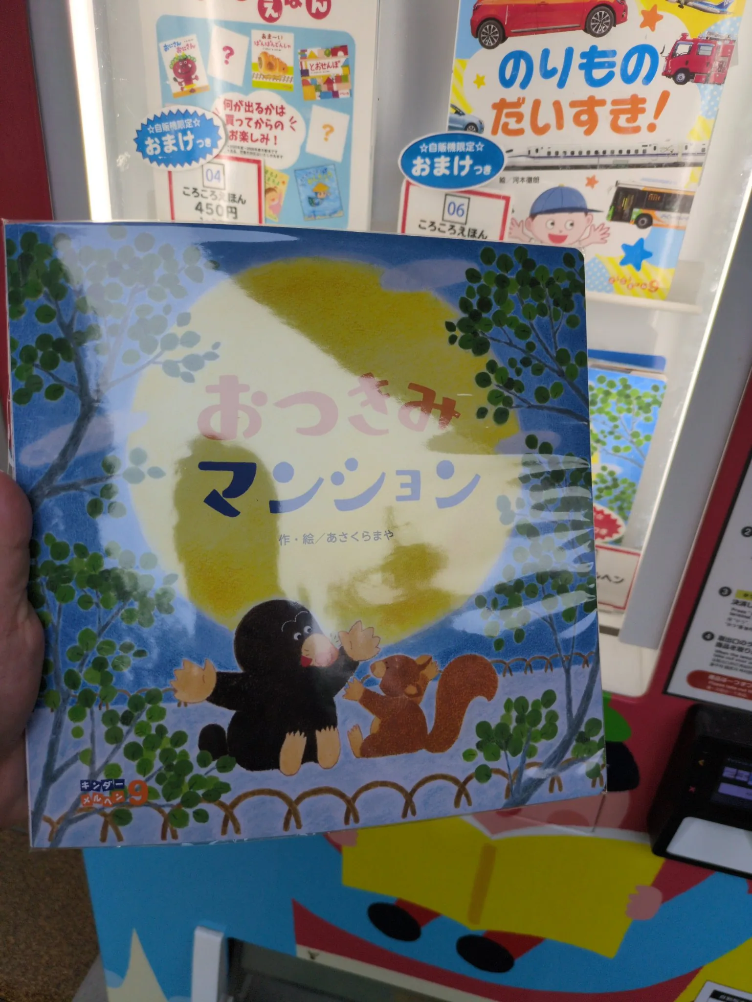 自販機の前に掲げた『おつきみマンション』の表紙。満月を背に、下辺の弓なりの柵に腰かけたモグラとリスが手を上げて向かい合っている。透明フィルムに包まれたまま。奥の棚に別の絵本が並ぶ